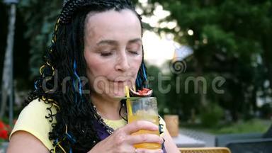 高级奢侈的女人喝着一杯激情水果鸡尾酒，坐在公园里的一家街头咖啡馆里，在树上。